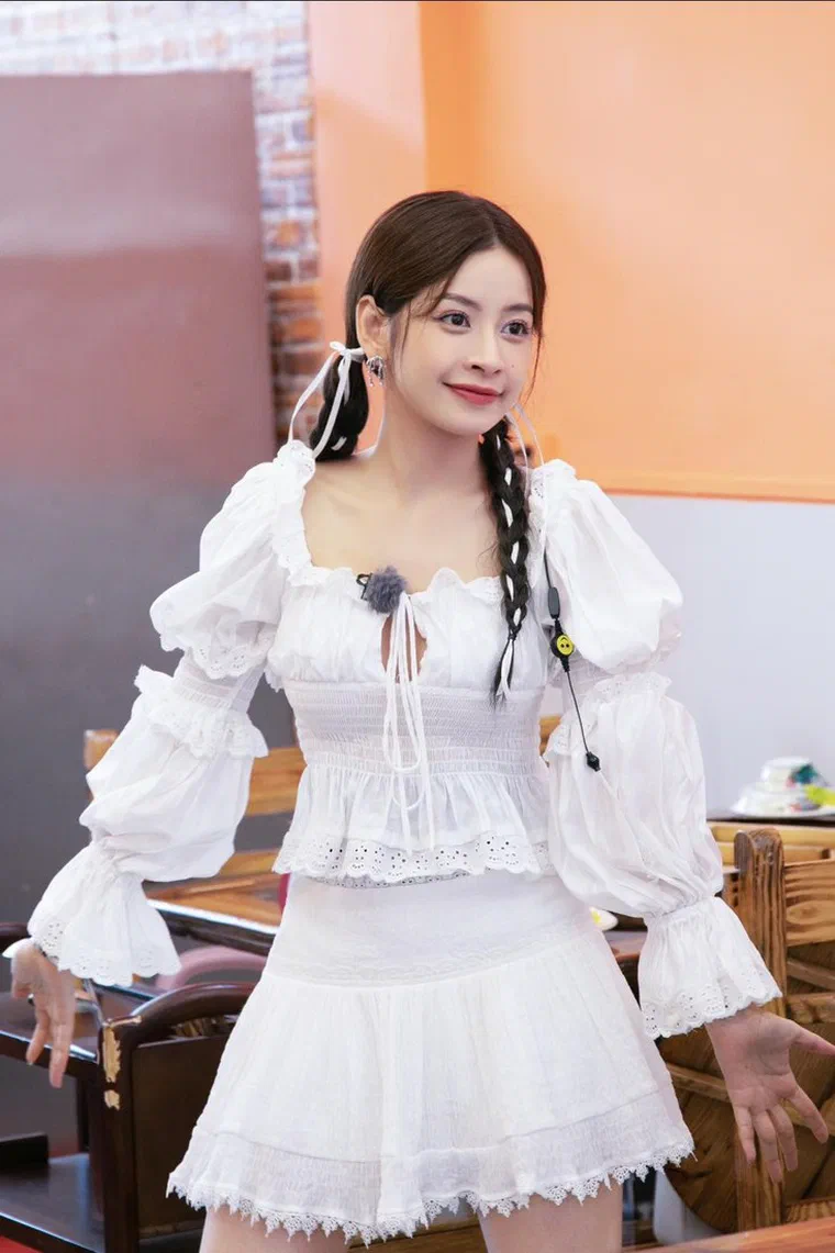 Chi Pu tóc nâu sẫm rẽ ngôi giữa, tết hai bím thấp sau tai buộc ruy băng trắng, vài sợi tóc mảnh rủ trước trán, mặc áo bèo trắng tay bồng