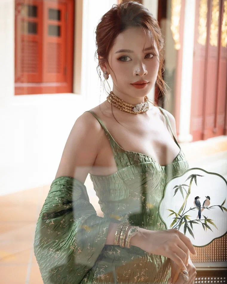 Chi Pu tóc nâu ánh đỏ vấn búi thấp lơi phía sau, đỉnh chải phồng nhẹ, hai lọn tóc mai uốn rủ cạnh má, đeo vòng cổ vàng, mặc váy corset xanh ngọc