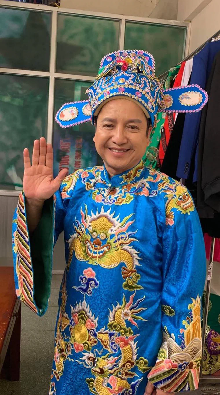Chí Trung mặc áo bào xanh lam thêu rồng vàng, đội mũ Táo xanh lam đính đá, giơ tay chào trong phòng phục trang, chỉ lộ tóc mai đen ngắn