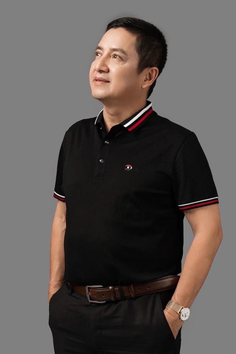 Chí Trung mặc áo polo đen viền cổ đỏ trắng, đứng nghiêng nhìn lên, tóc đen ngắn rẽ ngôi lệch, phần mái chải ngược gọn ra sau