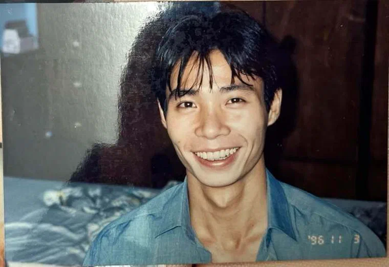Công Lý thời trẻ mặc sơ mi xanh, cười tươi, tóc đen dày để dài chấm gáy, phần mái rủ lòa xòa trước trán