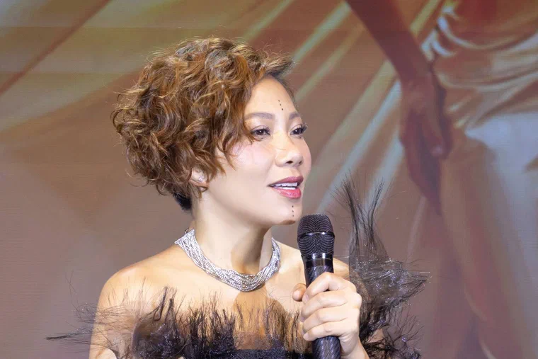 Hà Trần tóc xoăn lọn nhỏ ngắn, nền nâu sáng xen các lọn ánh vàng, phần đỉnh phồng, gáy cắt ngắn, cầm micro