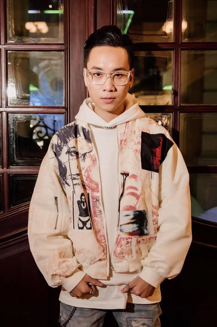 JustaTee đeo kính gọng trong, tóc ngắn chải gọn khỏi trán, mặc hoodie kem và áo khoác hoạ tiết