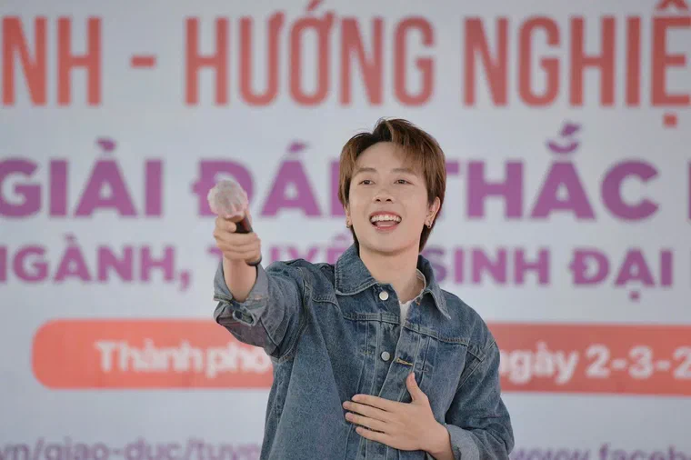 Nicky tóc nâu mật ong ngọn sáng hơn, rẽ ngôi giữa, mái chia hai bên, mặc áo khoác jean, cầm micro chỉ về phía khán giả