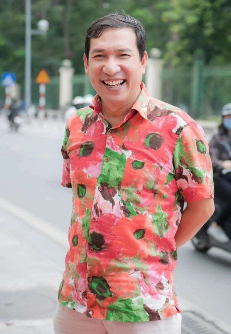 Quang Thắng mặc sơ mi hoa đỏ xanh, cười lớn trên phố, tóc đen rẽ ngôi lệch, phần đỉnh dài hơn và để rối tự nhiên