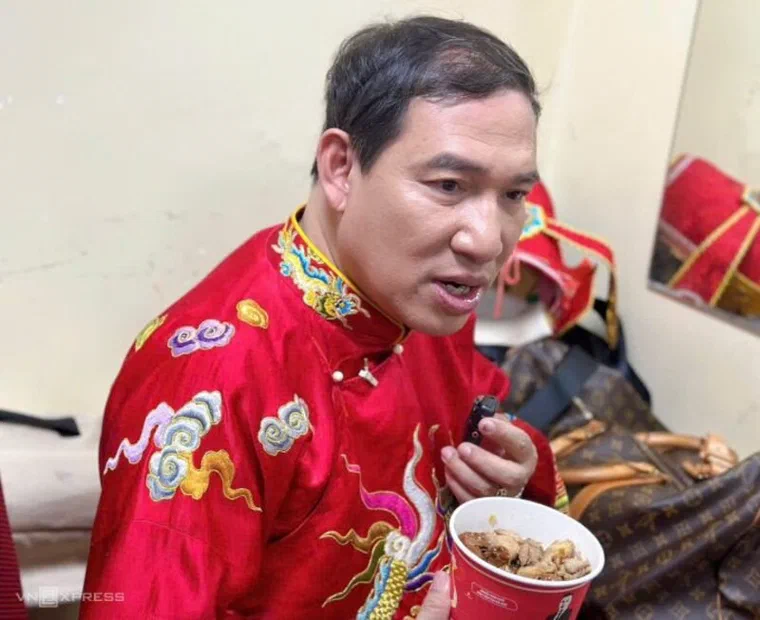 Quang Thắng mặc áo bào đỏ thêu rồng trong phòng chờ, tóc đen ngắn hai bên, phần đỉnh vuốt ngược và phồng nhẹ
