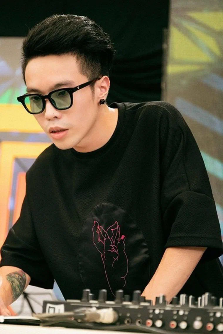 Triple D đeo kính râm đứng sau bàn DJ, hai bên đầu cạo sát, phần đỉnh để dài vuốt lệch về sau