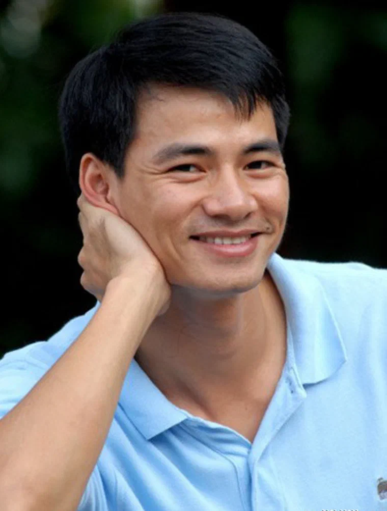 Xuân Bắc mặc áo polo xanh nhạt, tay chống cằm, tóc đen ngắn chải xuôi sang một bên, hai bên mai cắt gọn trên tai