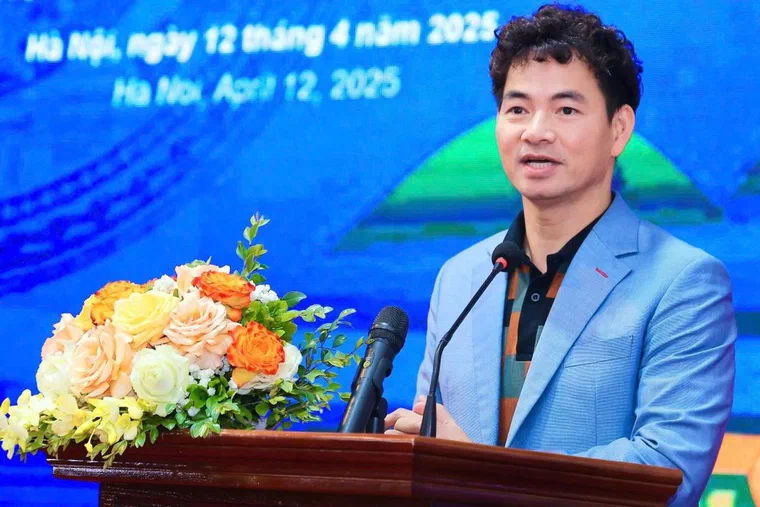 Xuân Bắc mặc vest xanh nhạt, phát biểu sau bục có hoa, tóc đen uốn xoăn lọn nhỏ phủ kín phần đỉnh, hai bên mai ngắn